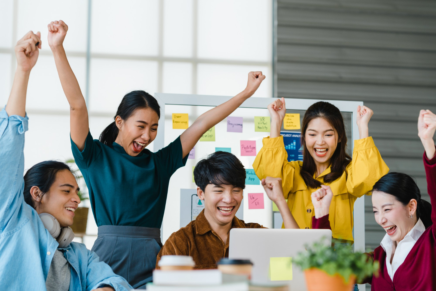 Diverse team of young professionals celebrating success with raised fists and big smiles in a modern office environment, representing successful recruitment outcomes and team building